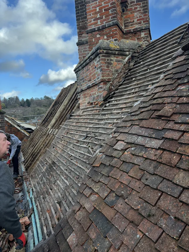 Old reddish tiles remain beside exposed battens and a tall brick chimney.