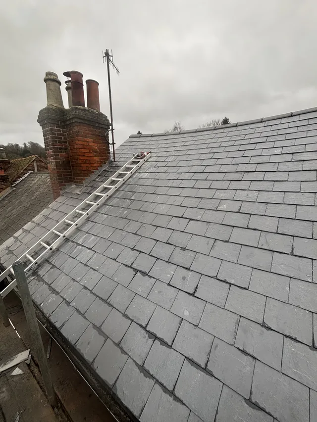 A roof ladder rests across grey slates beside a brick chimney with three pots.