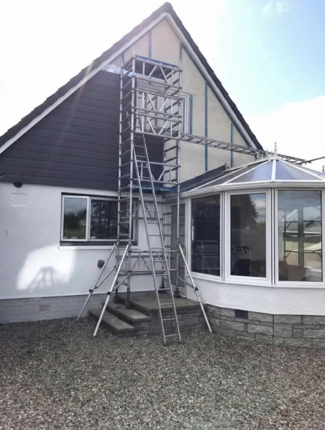 Dark horizontal cladding partly covers a gable behind a scaffold tower and white conservatory.