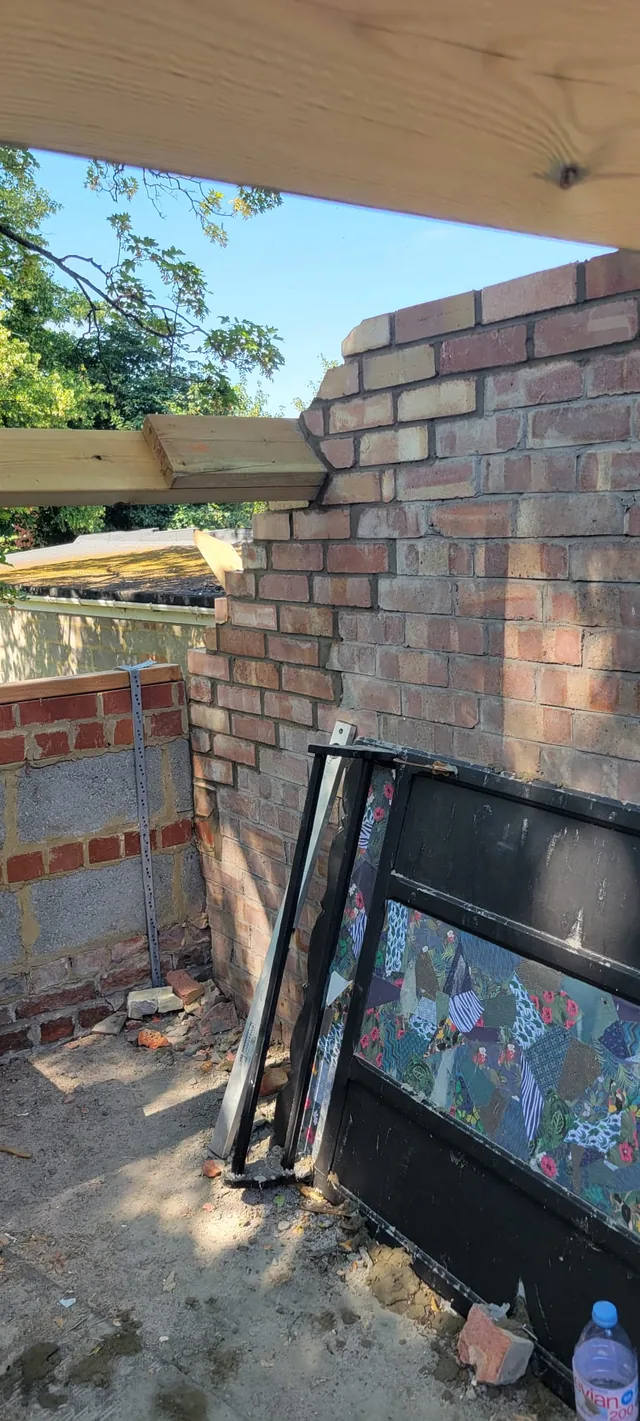 New brick courses rise beneath a timber beam with stored doors leaning below.