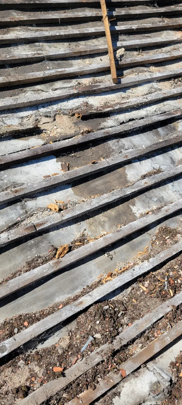 Debris and crumbling material sit between weathered roof battens beside a wooden ladder.