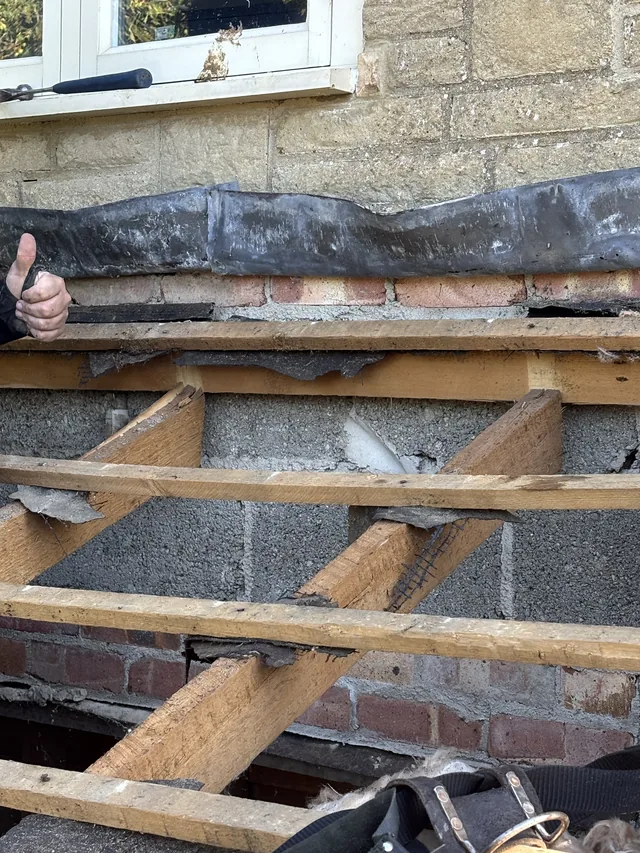 Exposed porch joists meet a stone wall beneath old flashing, with a hand at the edge.