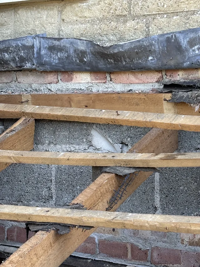 Timber porch joists span blockwork beneath weathered flashing along a stone wall.
