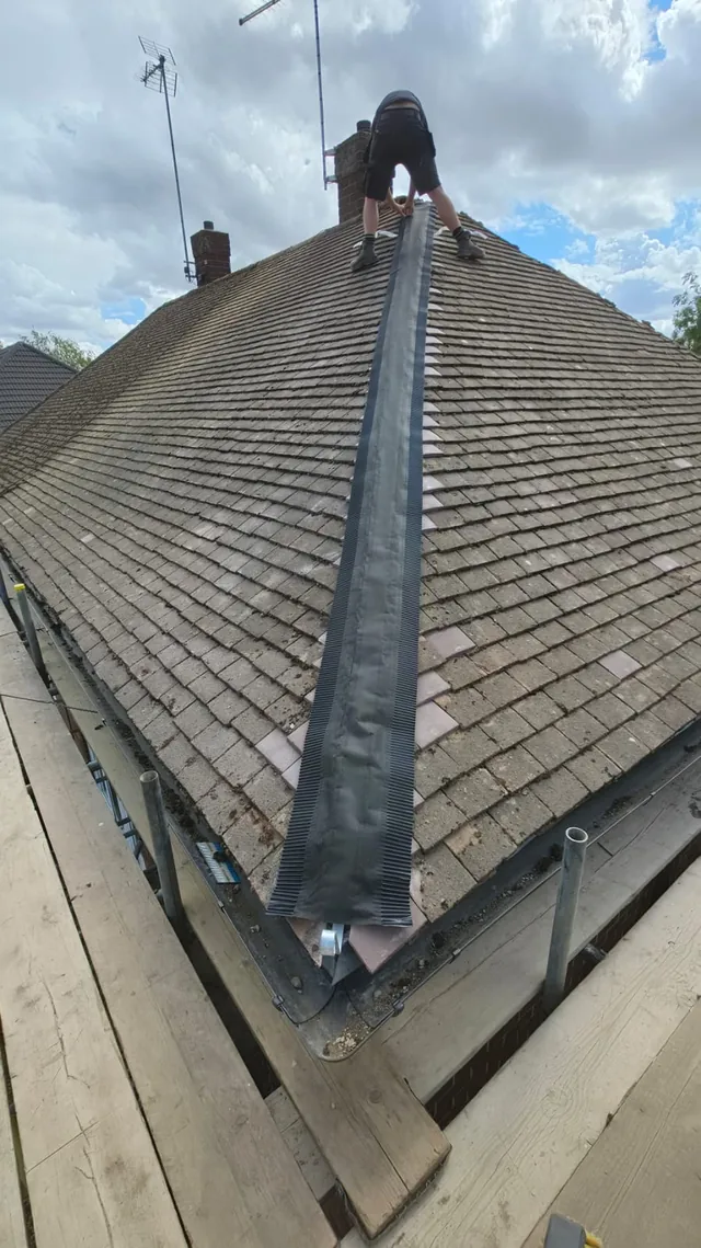 A roofer works at the top of dark hip roll between two tiled slopes.