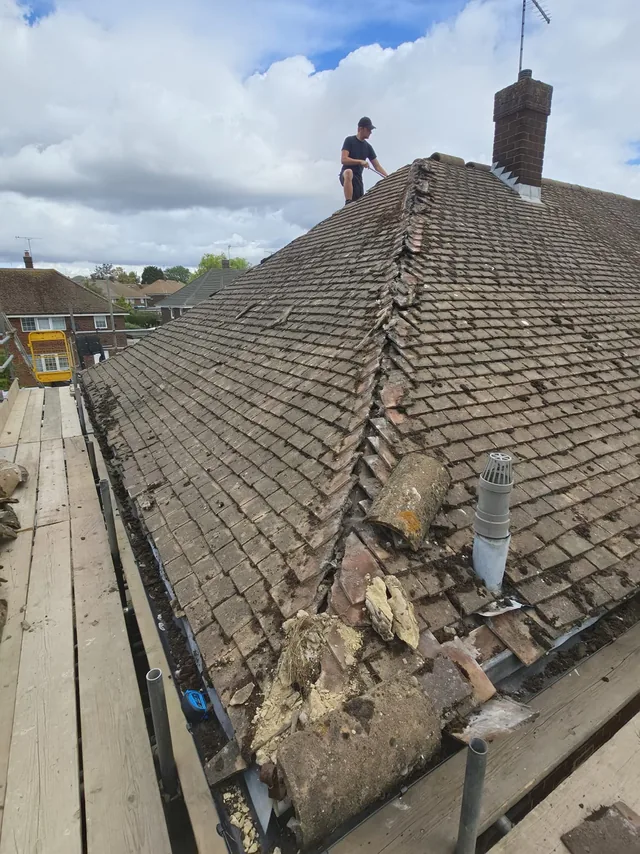 A roofer stands above an opened hip with broken mortar and a nearby flue.