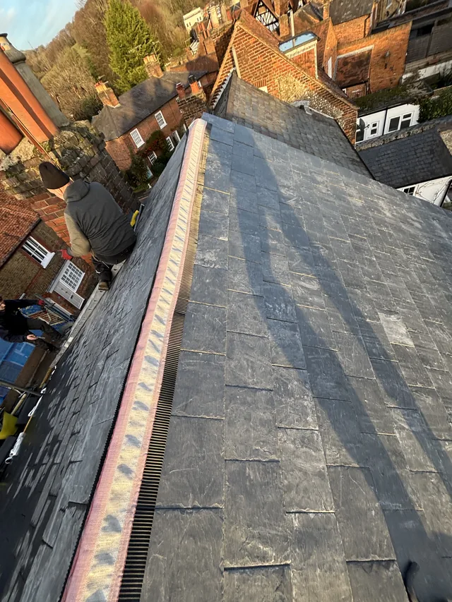 A strip of ridge material runs between slate slopes beside roofers and scaffolding.