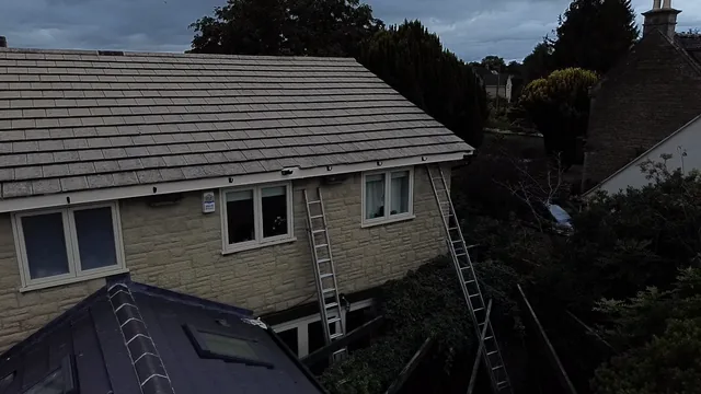 Dark guttering follows the tiled eaves above white windows on a stone house.