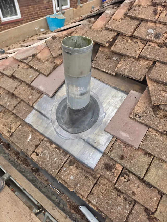 A new lead slate surrounds an open flue pipe beside displaced brown roof tiles.