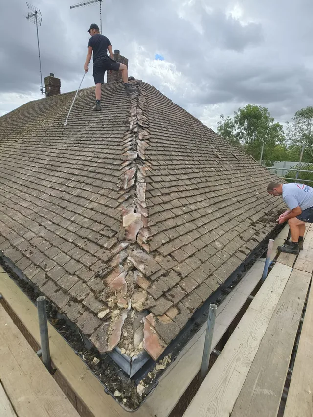Two roofers work beside an opened hip with loose mortar and weathered roof tiles.