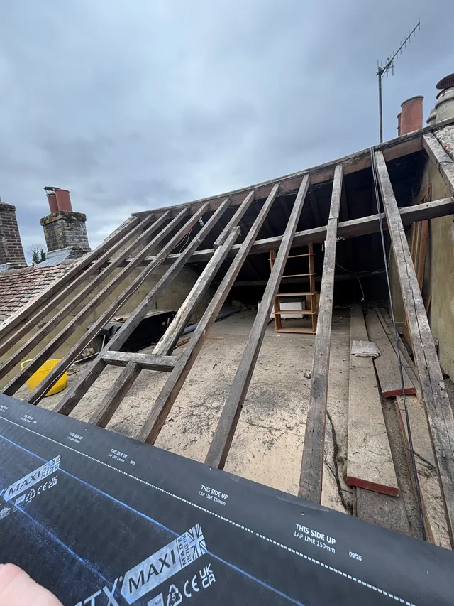 Bare roof rafters and interior boards rise above a roll of dark membrane.