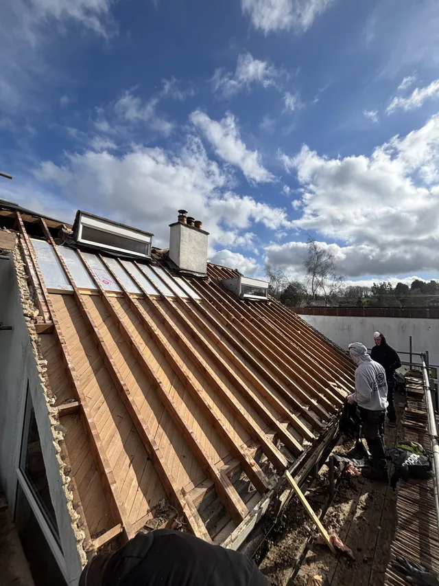 Two roofers stand beside exposed rafters and white panels beneath scattered clouds.