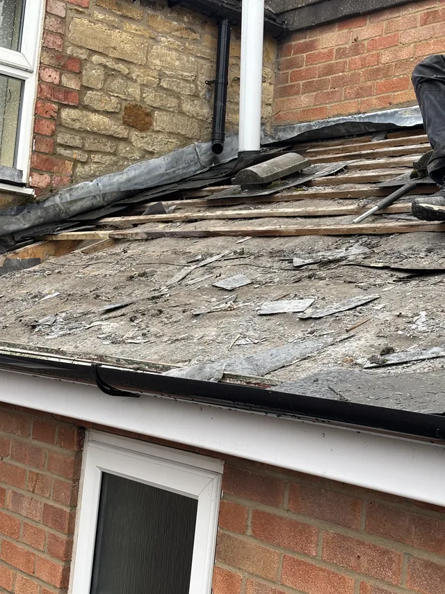 Loose slate fragments lie across a stripped extension roof below white drainage pipes.