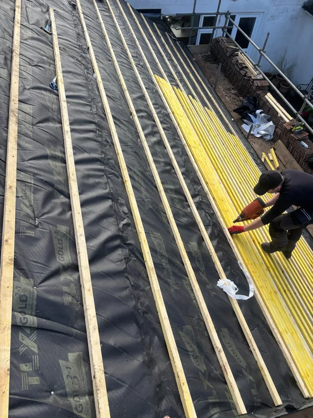 A roofer kneels over yellow battens beside dark membrane on a steep roof slope.