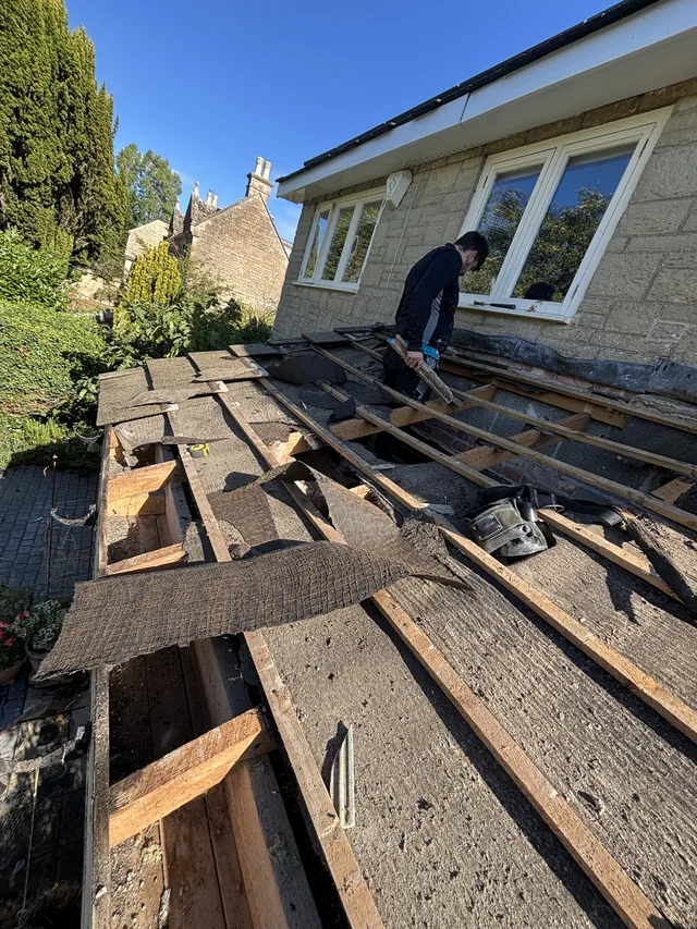 A roofer strips a porch roof to timber framing beneath stone walls and windows.