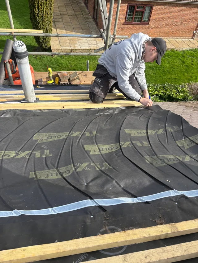 A kneeling roofer fits dark membrane beside a flue and scaffold rails.
