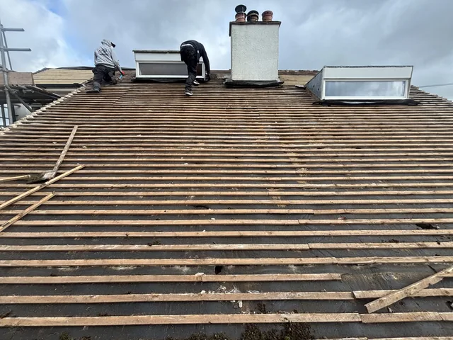 Two roofers work near rooflights above a wide slope of exposed battens.