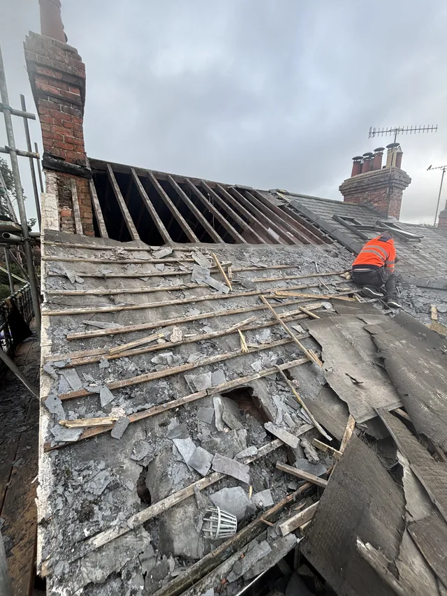 A worker in orange strips an old roof between two brick chimney stacks.