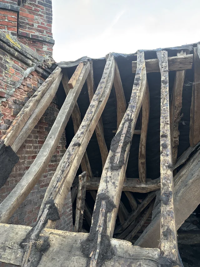 Weathered roof rafters stand exposed beside a brick chimney beneath an overcast sky.