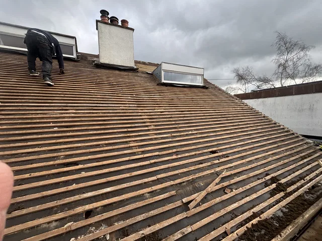 A roofer bends over bare battens beneath two rooflights and a rendered chimney.