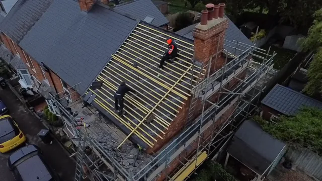 Two roofers work across yellow battens beside a brick chimney and surrounding scaffolding.