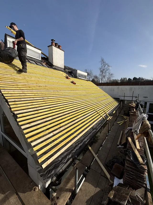 Roofers work across a newly battened slope beneath rooflights and a white chimney.