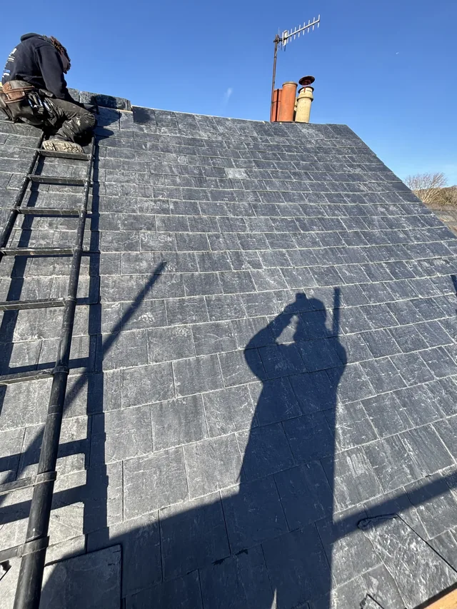 A roofer works near the ridge above grey slates and a roof ladder.