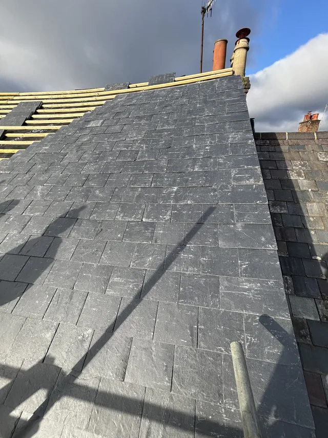 Grey slates climb towards exposed battens near a chimney, crossed by scaffold shadows.