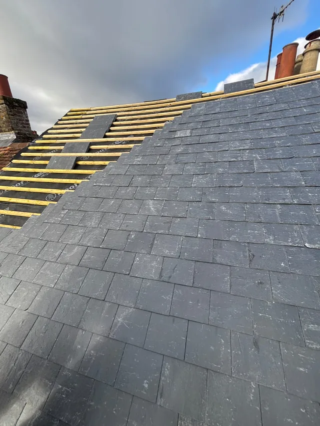 Grey slate courses rise towards chimney pots while yellow battens remain exposed near the ridge.