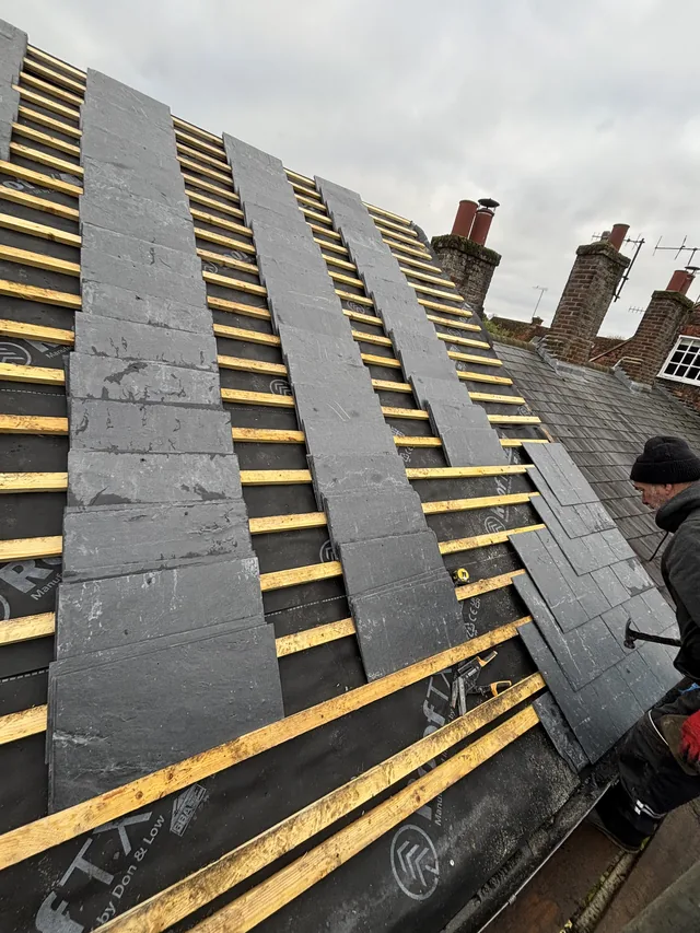 Vertical runs of grey slates rise across yellow battens beside a crouching roofer.