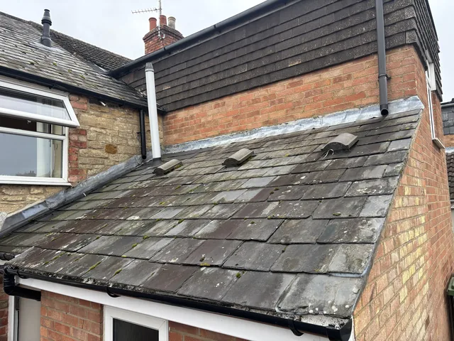 Weathered slates cover an extension beneath brickwork, dark wall tiles and white drainage pipes.
