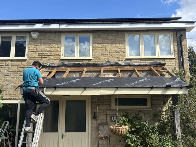 A roofer on a ladder fits dark membrane across a porch beneath upstairs windows.