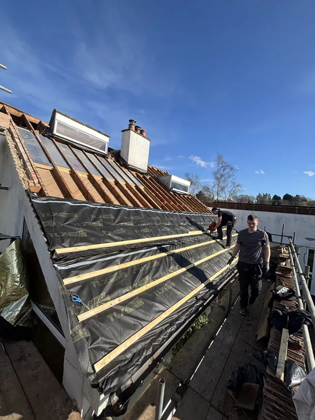 Roofers stand on scaffolding beside newly exposed rafters and partly fitted dark membrane.