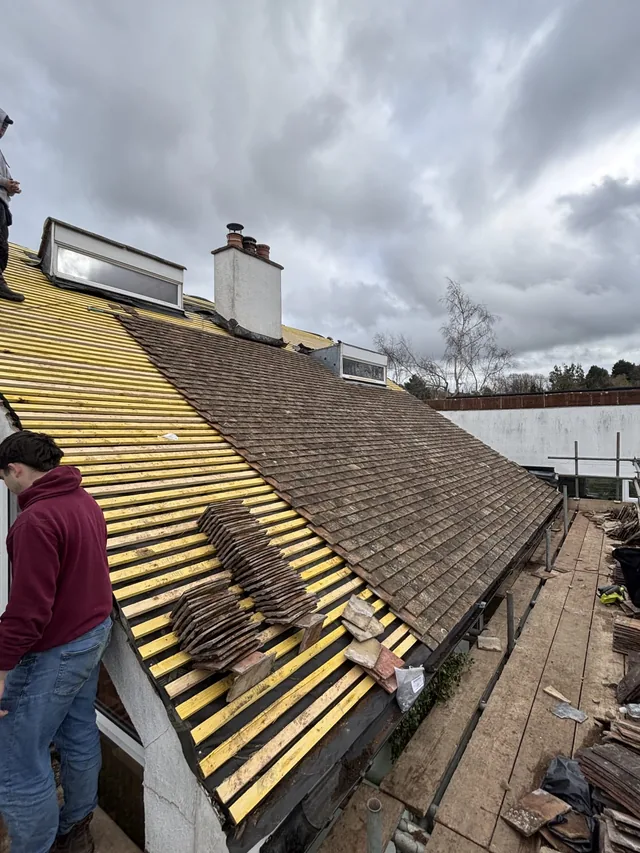 Stacks of brown tiles sit on yellow battens beside a partly covered roof slope.