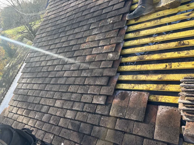 Weathered brown tiles meet exposed yellow battens, with stacked spare tiles beside them.